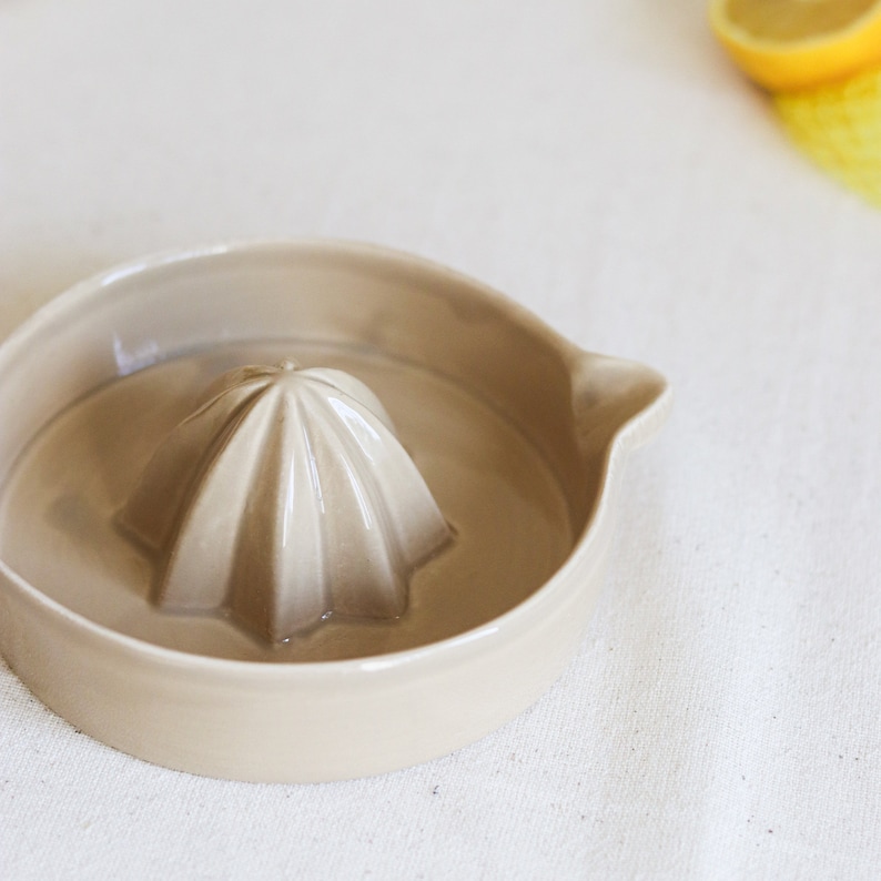 May include: A beige ceramic citrus juicer with a fluted reamer in the center. The juicer is on a white linen surface.