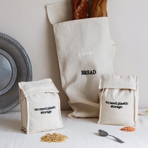 Könnte beinhalten: Drei weiße Stoffbeutel mit schwarzem Sticktext. Der größte Beutel sagt "BREAD" und die beiden kleineren Beutel sagen "no need plastic storage."