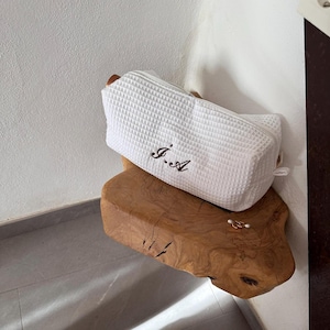 Può includere: Un beauty case rettangolare bianco con trama a nido d'ape, appoggiato su una superficie in legno. La borsa presenta le iniziali ricamate "J.A" in marrone scuro. La borsa ha una chiusura a zip e un piccolo anello laterale.