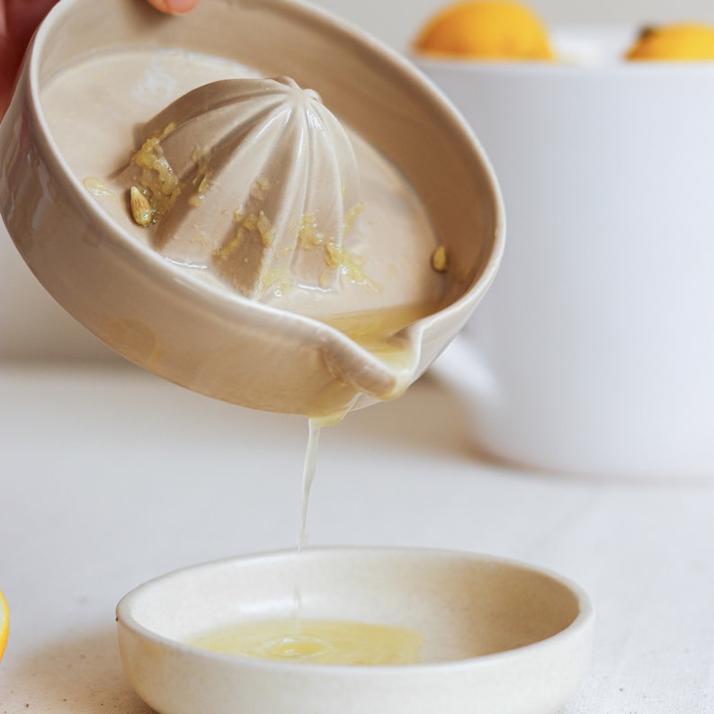 May include: A ceramic lemon squeezer with a light brown glaze is being used to squeeze juice into a small, white ceramic bowl. The bowl is sitting on a white surface.