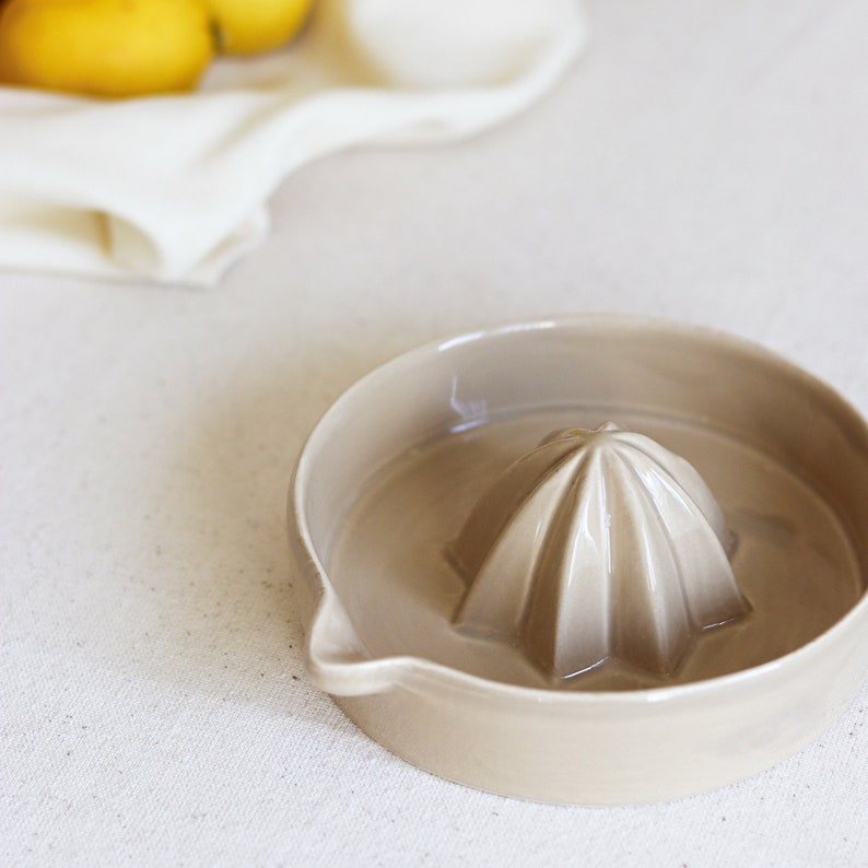 May include: A beige ceramic citrus juicer with a ridged reamer in the center. The juicer has a spout for pouring juice.