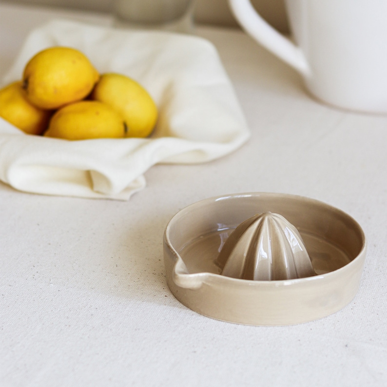 May include: A beige ceramic lemon squeezer with a ridged design. The squeezer is on a white linen surface.