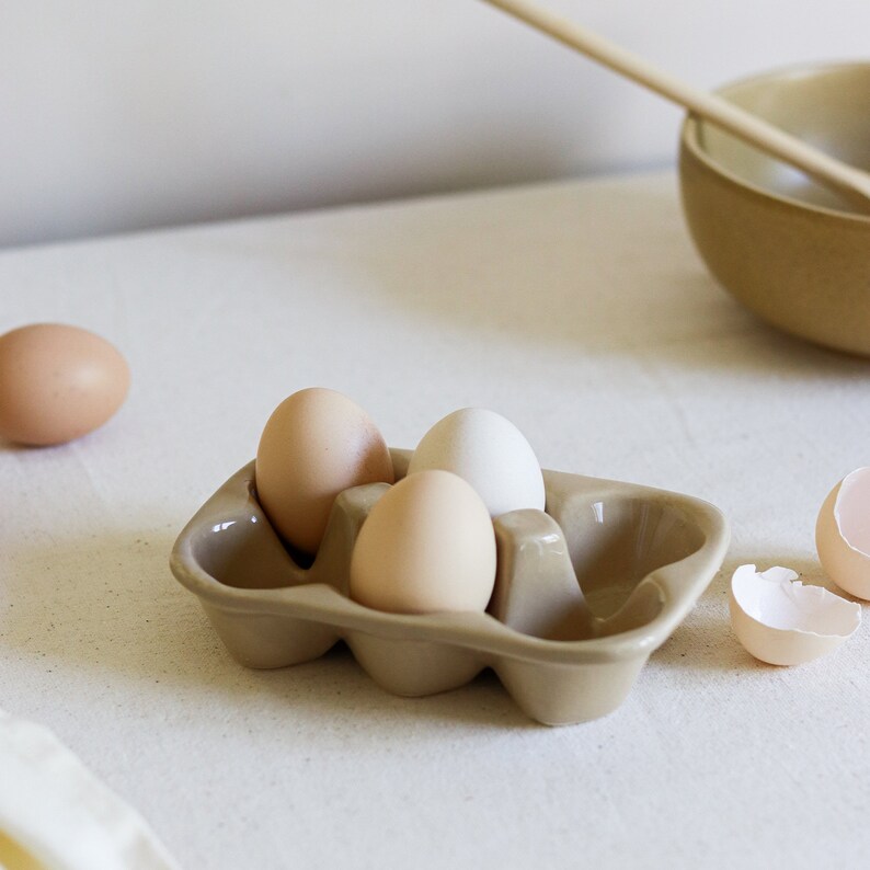 May include: A beige ceramic egg holder with four eggs inside. Three of the eggs are brown and one is white. The holder is sitting on a white surface.