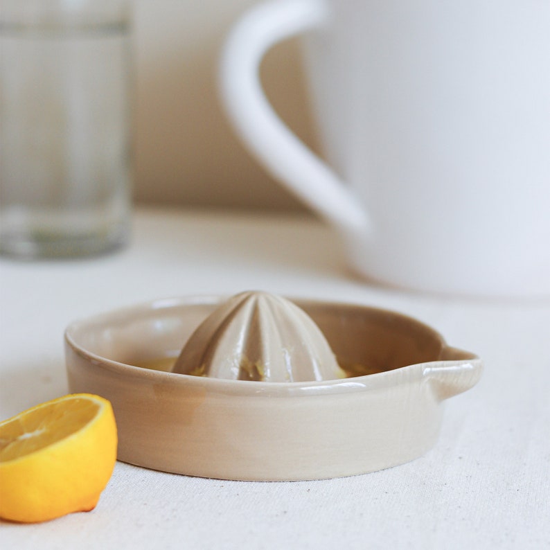 May include: A beige ceramic lemon squeezer with a ridged top and a small spout. The squeezer is sitting on a white surface with a lemon wedge next to it.