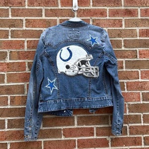 Puede incluir: Chaqueta vaquera azul con un diseño de casco de fútbol americano blanco y plateado y detalles de estrellas azules. La chaqueta está colgada de un gancho blanco contra una pared de ladrillos. La chaqueta presenta el logotipo de los Colts de Indianápolis.