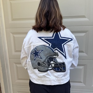 May include: White denim jacket with a large navy blue star and a football helmet graphic on the back. The jacket features a classic design with a button-up front and a collared neckline. The jacket is a fan item.