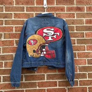 May include: A blue denim jacket with a large patch featuring the San Francisco 49ers logo and a football helmet. The logo is red and gold. The jacket is hanging on a white hook against a brick wall.