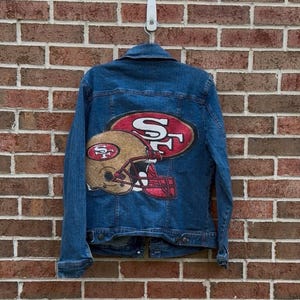 Könnte beinhalten: Blaue Jeansjacke mit einem großen, farbenfrohen Aufdruck eines Footballhelms und den Buchstaben "SF" auf dem Rücken. Die Jacke hängt vor einer Backsteinmauer.