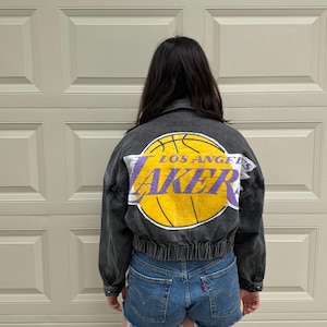May include: A black denim jacket with a Los Angeles Lakers logo on the back. The logo features a yellow basketball with the words "LOS ANGELES" and "LAKER" in purple. The jacket has a ribbed hem and cuffs.