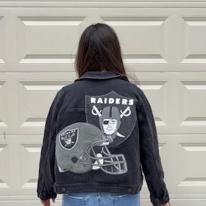 Könnte beinhalten: Eine schwarze Jeansjacke mit einer großen Grafik auf der Rückseite. Das Design zeigt einen Football-Helm und ein schildförmiges Logo mit dem Schriftzug RAIDERS. Die Jacke hat lange Ärmel, einen Kragen und Knopfverschlüsse und wird über einer blauen Jeans getragen.