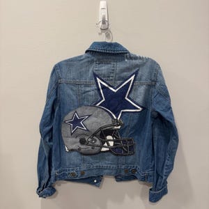 Könnte beinhalten: Eine blaue Jeansjacke mit einem großen marineblauen Stern und einem Footballhelm-Patch. Die Jacke hat eine Knopfleiste, einen Kragen und lange Ärmel. Die Aufnäher sind in den Farben des Teams.