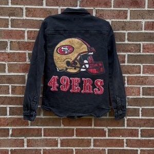 May include: Black denim jacket featuring a gold and red football helmet graphic and the text "49ERS" on the back. The jacket has long sleeves, a button-down front, and a classic collar. The jacket is hanging on a brick wall.