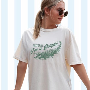 May include: A white t-shirt with a green alligator graphic and the text "First of all, I'm a Delight". The shirt is shown on a model and the background is a light blue and white striped pattern. The text "Ivory" is shown in the bottom right corner of the image.