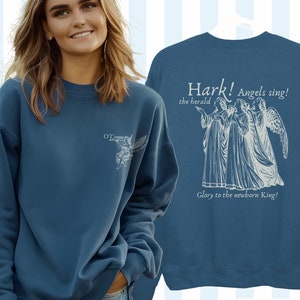 May include: Indigo blue sweatshirt with a white angel graphic and the text "Hark! Angels sing! the herald Glory to the newborn King!"