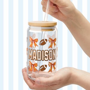 May include: A clear glass tumbler with a wooden lid and a straw. The tumbler has a design of orange bows and brown footballs. The name "MADISON" is printed in orange on the tumbler.