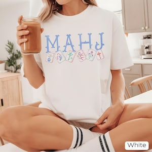 May include: White t-shirt with the word "MAHJ" in light blue letters above a row of floral-themed playing cards. The person is holding a glass of brown liquid. The shirt is paired with white socks with black stripes.