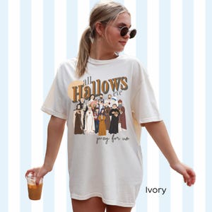 K&ouml;nnte beinhalten: Ein wei&szlig;es T-Shirt mit einem grafischen Druck einer Gruppe von Personen in religi&ouml;ser Kleidung. Der Text "all Hallows eve pray for us" ist auf das T-Shirt gedruckt. Das T-Shirt ist mit "Ivory" gekennzeichnet.