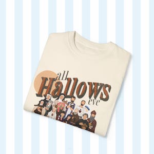 K&ouml;nnte beinhalten: Ein beiges T-Shirt mit einem braunen Grafikdruck, auf dem "all hallows eve" steht, mit einer Gruppe von Personen im Hintergrund.