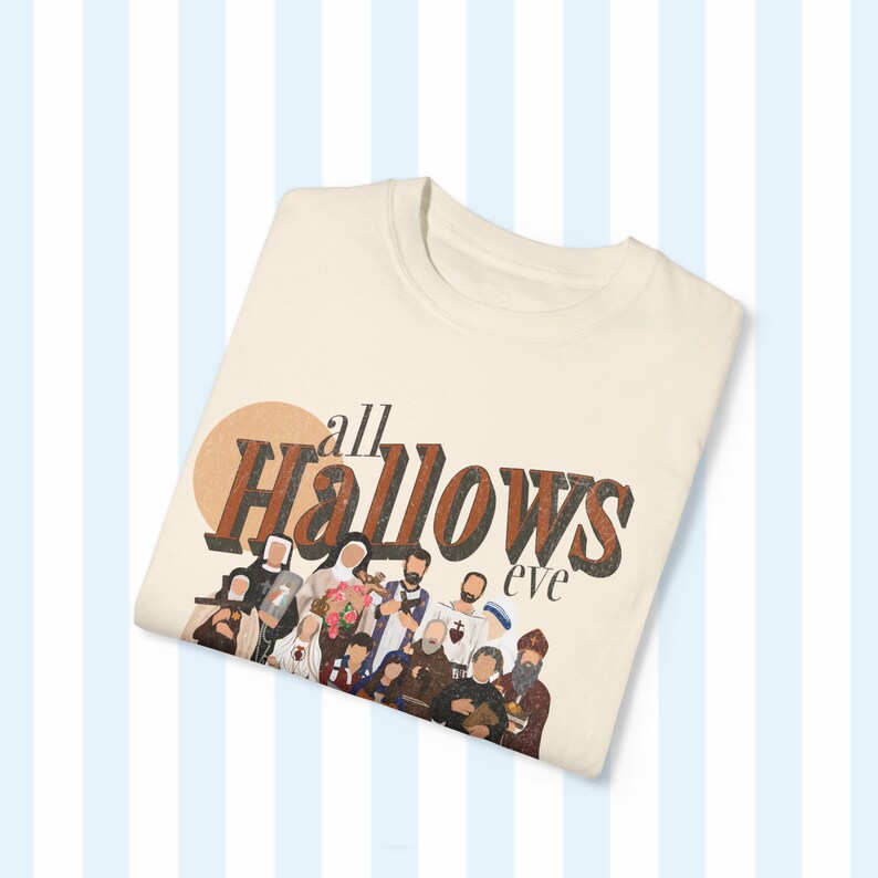 K&ouml;nnte beinhalten: Ein beiges T-Shirt mit einem braunen Grafikdruck, auf dem "all hallows eve" steht, mit einer Gruppe von Personen im Hintergrund.