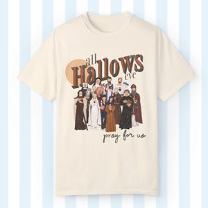 K&ouml;nnte beinhalten: Ein beiges T-Shirt mit einer Grafik im Vintage-Stil einer Gruppe von Personen in religi&ouml;ser Kleidung. Die Grafik lautet "all Hallows eve pray for us".