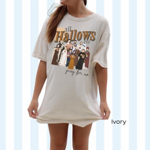 K&ouml;nnte beinhalten: Ein T-Shirt in Elfenbein mit einem grafischen Druck einer Gruppe von Personen in religi&ouml;ser Kleidung. Der Text "all hallows eve pray for us" ist auf das T-Shirt gedruckt.