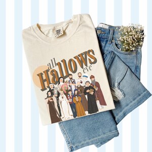 K&ouml;nnte beinhalten: Ein beiges T-Shirt mit dem Text "all Hallows eve" in braunen und schwarzen Buchstaben. Das Shirt zeigt eine Grafik einer Gruppe von Menschen in religi&ouml;ser Kleidung.