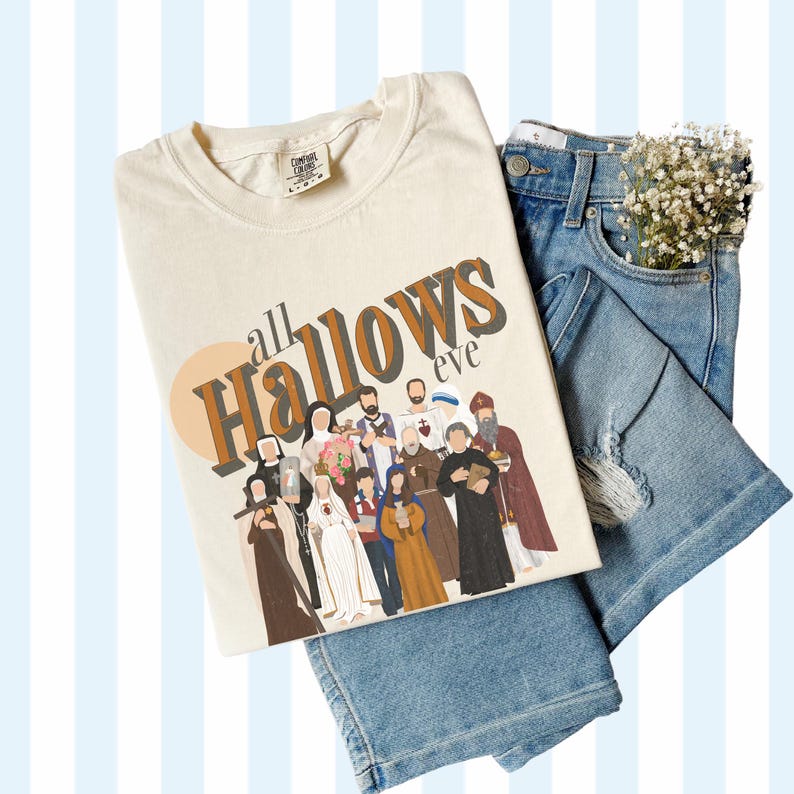 K&ouml;nnte beinhalten: Ein beiges T-Shirt mit dem Text "all Hallows eve" in braunen und schwarzen Buchstaben. Das Shirt zeigt eine Grafik einer Gruppe von Menschen in religi&ouml;ser Kleidung.