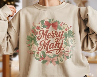 May include: Sand-colored sweatshirt featuring a festive wreath design with the words "Merry Mahj" in a script font. The wreath includes red bows, holly, and Mahjong tiles. The word "Sand" is printed at the bottom.