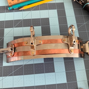 May include: A curved metal cuff featuring alternating horizontal bands of silver-toned and copper-toned metal. The piece is secured with small brass screws and includes three swivel snap hooks attached to the front, with a D-ring on the side.