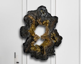 Op de afbeelding: Een abstract wandkunstwerk in zwart en goud met een unieke organische vorm. Het kunstwerk hangt aan een witte muur boven een bruine fauteuil en een zwarte bijzettafel.