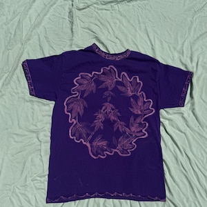 May include: A short-sleeved purple t-shirt featuring a large, central graphic of a peace sign composed of detailed leaf patterns in a lighter shade of purple. The shirt includes decorative, intricate trim along the neckline, sleeve edges, and bottom hem.