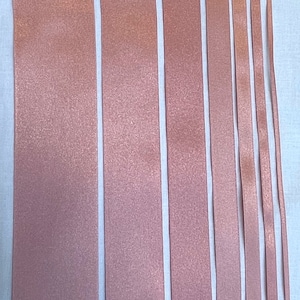 May include: Assortment of seven different widths of rose gold satin ribbon. The ribbon is arranged vertically on a white surface.