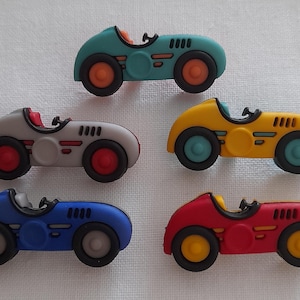 May include: Five toy race cars in various colours: teal, grey, yellow, blue, and red. Each car features black wheels with coloured accents, a driver's seat, and an open-top design. The cars are displayed on a white surface.