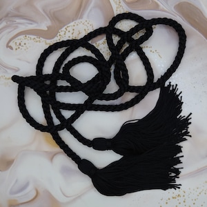 May include: Black rope with two large tassels. The rope is knotted and looped, with a thick, braided texture. The tassels are made of many strands of black material, creating a full, soft appearance. The background is a marbled pattern with gold flecks.