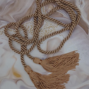 May include: A decorative cord with two tassels. The cord is a light brown colour, with a braided design. The tassels are made of a similar material and colour, adding a touch of elegance. The cord is likely used for decorative purposes.