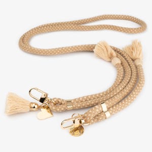 Puede incluir: Una correa de perro de cuerda beige con lunares blancos y herrajes dorados. La correa tiene dos borlas en cada extremo y un dije en forma de corazón.