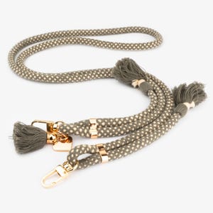 Puede incluir: Un cordón de cuerda con estampado verde y blanco con herrajes metálicos dorados. El cordón tiene una borla en cada extremo y un dije en forma de corazón. La cuerda es gruesa y parece estar hecha de un material duradero.