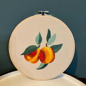 May include: Embroidered hoop art featuring two peaches with green leaves. The peaches are a vibrant orange and red color.