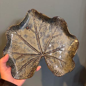 May include: A ceramic leaf-shaped dish with a textured surface and a glossy finish. The dish features a central vein pattern and is primarily shades of brown and blue. The edges are slightly wavy, adding to the natural aesthetic.