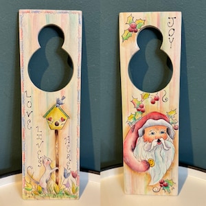 Könnte beinhalten: Zwei dekorative Türanhänger aus Holz. Einer zeigt eine Katze, ein Vogelhaus und den Text "LOVE LIVES HERE". Der andere zeigt den Weihnachtsmann mit Stechpalme und dem Wort "JOY". Beide haben einen achtförmigen Ausschnitt.