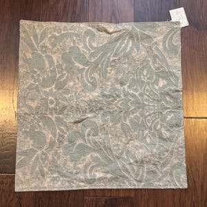 May include: A square throw pillow cover featuring a light sage green and off-white damask pattern. The fabric displays an intricate, woven floral and scrollwork design across the entire surface. A small white rectangular product tag is attached.