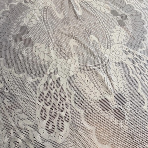 May include: Cream-colored lace tablecloth featuring a detailed peacock design. The intricate pattern includes peacock figures, feathers, and floral elements. The lace is delicate and adds a vintage aesthetic to the table setting.