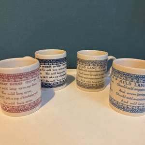 Puede incluir: Cuatro tazas de cerámica con asas, cada una con un diseño diferente. Las tazas tienen texto del poema "Auld Lang Syne" y "Robert Burns" en azul y rojo. Las tazas están sobre una bandeja blanca.