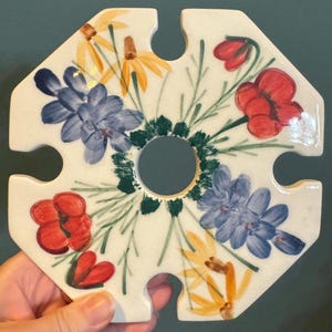 May include: A white ceramic trivet with a floral design. The trivet has a central hole and four cut-out sections. The flowers are painted in red, blue, and yellow, with green stems and leaves. The trivet is held by a hand.
