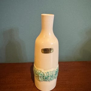 May include: A cream-colored ceramic vase with a narrow neck and a wider base. The vase features a band of geometric, turquoise-colored designs around the middle. The label reads "STUDIO SERIES".