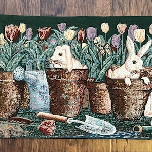 Puede incluir: Un tapiz que presenta una escena de jardín con conejos blancos asomándose de macetas llenas de tulipanes. También hay una regadera y herramientas de jardinería. El fondo es verde oscuro.