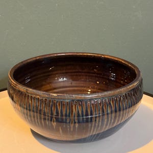 May include: A handmade ceramic bowl with a glossy finish. The bowl features a dark brown interior and a textured exterior with vertical lines and a band of blue and brown glaze. The bowl is a decorative piece for the kitchen or dining room.