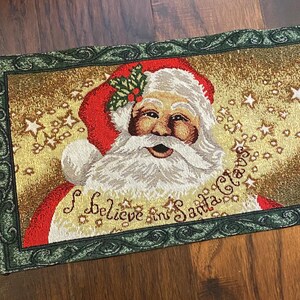 May include: A rectangular Christmas table runner featuring Santa Claus with a white beard and red hat. The text "I Believe in Santa Claus" is displayed below the image. The runner has a green border and a gold background with star accents.