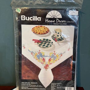 May include: A Bucilla Home Decor collection stamped cross stitch kit for a fruit garland tablecloth. The kit includes a printed fabric with a design of a tablecloth with a fruit garland border and a pie and teacups in the centre. The kit also includes DMC floss requirements available on instructions.
