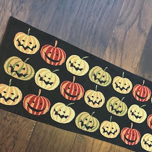 Puede incluir: Una alfombra rectangular de color negro con un patrón repetitivo de calabazas de Halloween en tonos naranja, amarillo y verde. Cada calabaza tiene una cara tallada con ojos triangulares y una boca dentada, dispuestas en una cuadrícula.